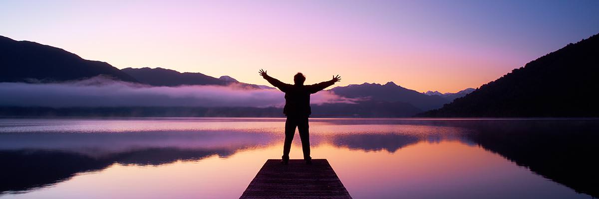 Person standing at the end of a dock looking across Lake Kaniere at sunrise, arms raised to greet the dawn