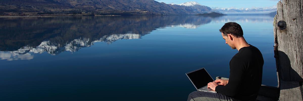 Man sitting alone at the edge of a peaceful lake working on his laptop