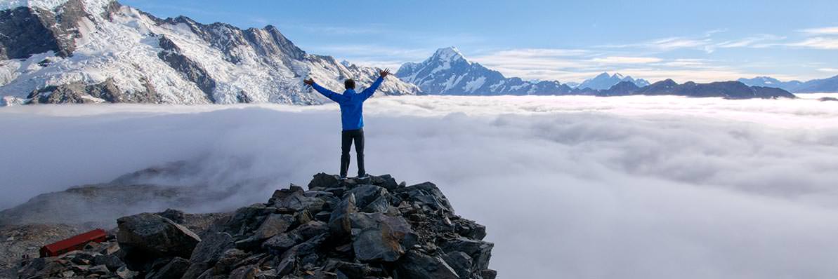 Person standing on a rocky outcrop above a mist-filled valley looking towards the Southern Alps, arms raised in triumph